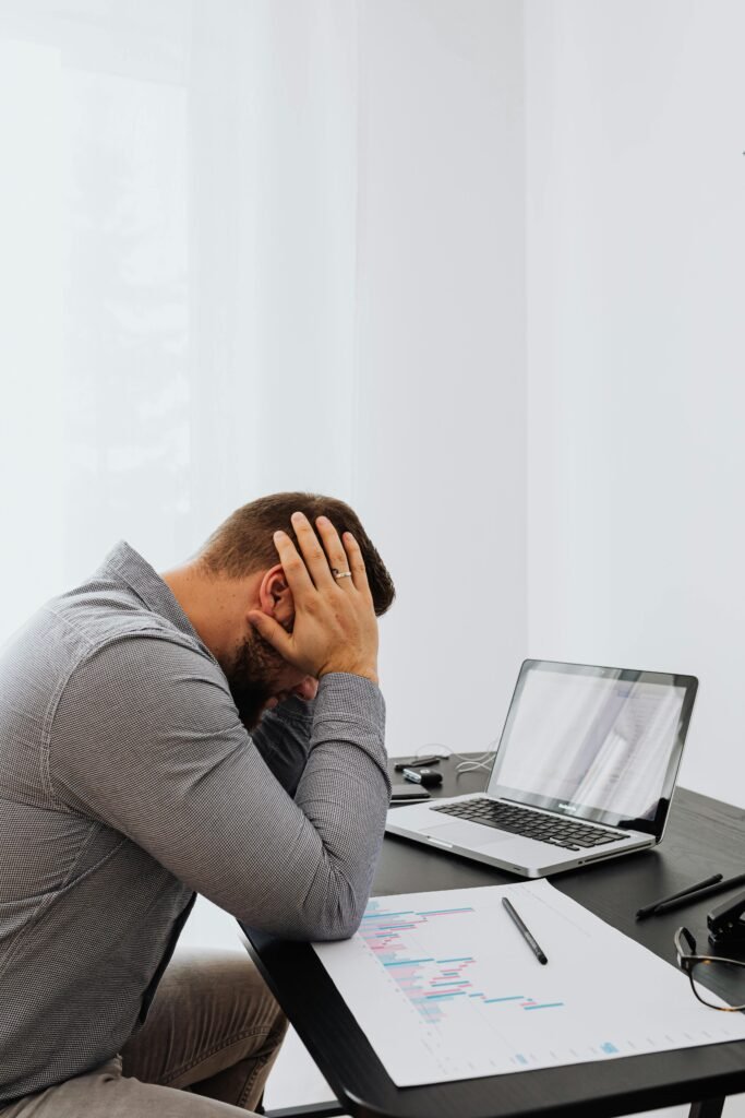 A frustrated man sits at a desk with a laptop and charts, showing work stress.